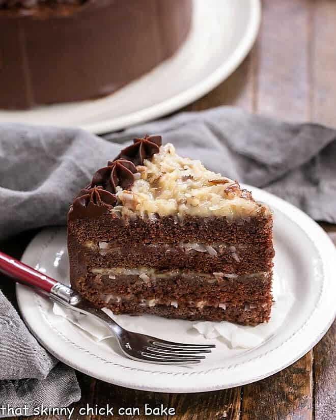 4 Layer Homemade Chocolate Cake filled German chocolate coconut icing - 4 layers of chocolate cake filled with a caramel coconut pecan filling, then iced with a rich, chocolate frosting! Perfect cake for any celebration! Slice of German Chocolate Cake on a white dessert plate in front of a partial view of the cake.