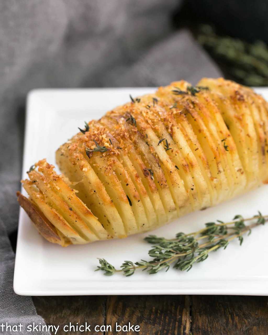 Hasselback Potatoes on a square white plate with a sprig of thyme.