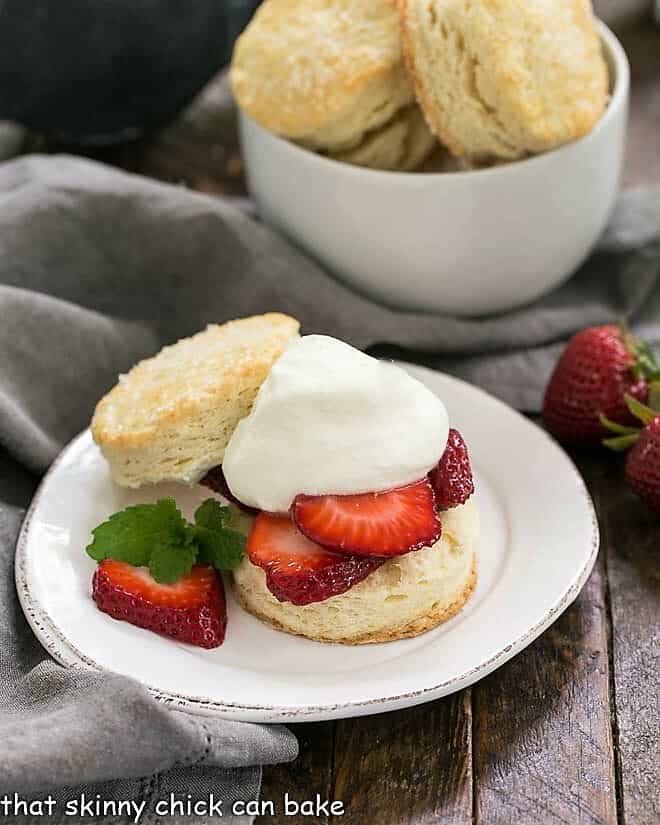 Classic Strawberry Shortcake - tender flaky biscuits topped with sweet, juicy berries and freshly whipped cream for a magnificent spring or summer dessert Classic strawberry shortcake on a plate in front of a bowl of shortcake biscuits.