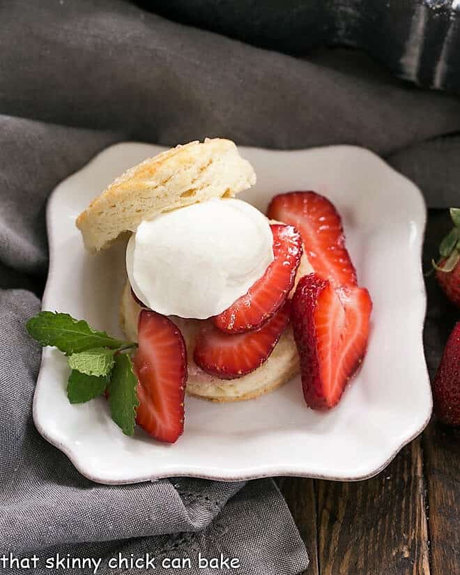Classic Strawberry Shortcake - tender flaky biscuits topped with sweet, juicy berries and freshly whipped cream for a magnificent spring or summer dessert Classic Strawberry Shortcake on a square white plate with a dollop of cream and mint sprig.