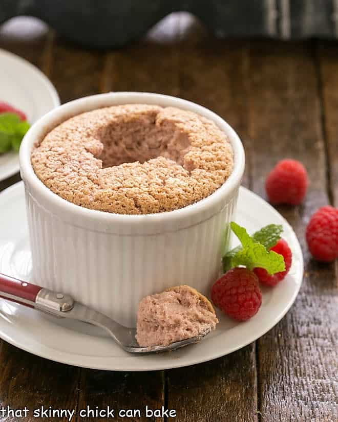 A raspberry souffle with a bite removed on a spoon in front of a ramekin