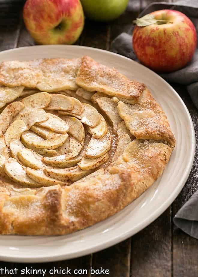 This scrumptious Apple Galette is a gorgeous, impressive dessert. This rustic apple tart has all the delicious flavors of a pie without all the effort! Rustic Apple Tart on a white plate with apples in the background.