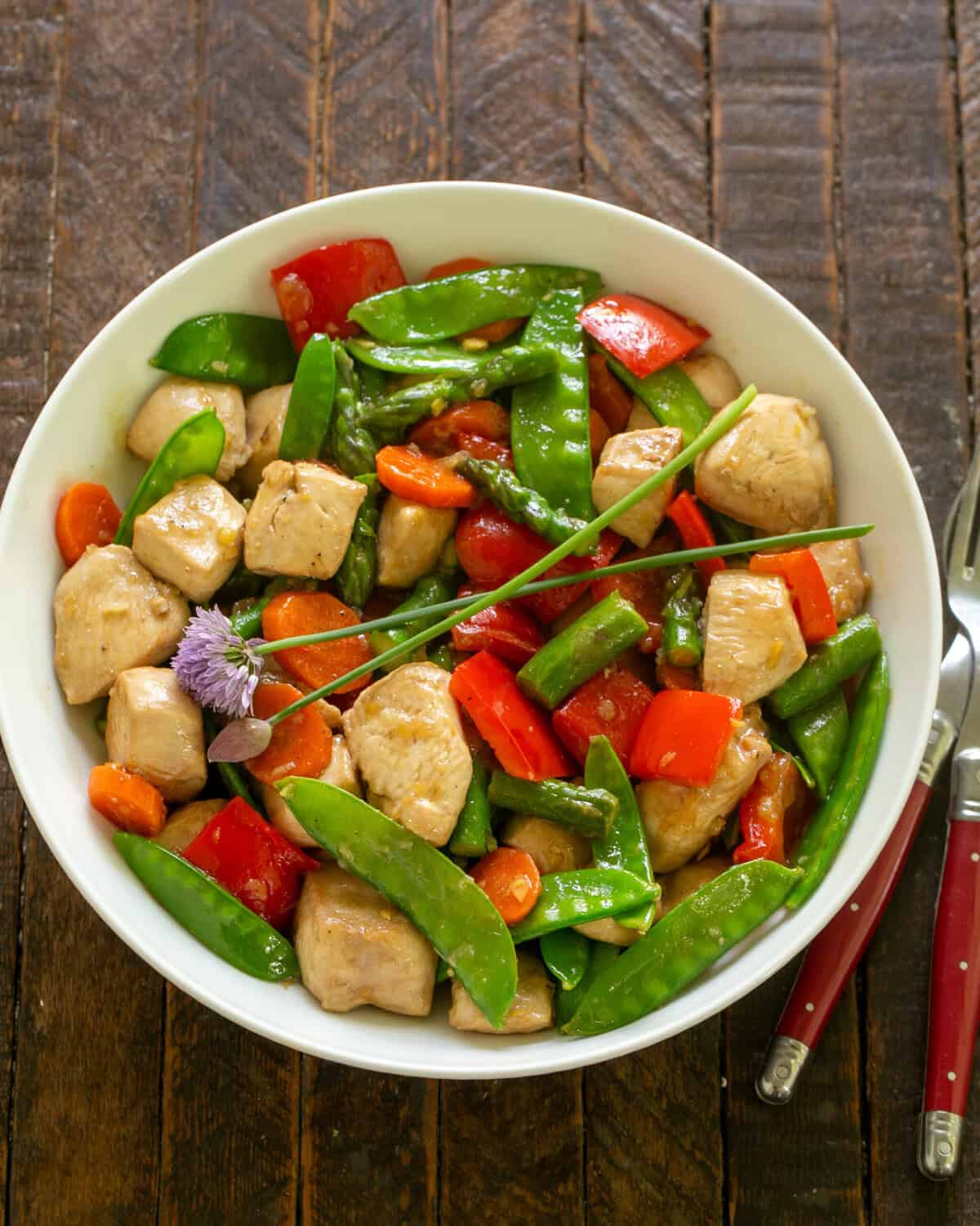 Chicken Stir Fry in a white serving bowl with red handled fork and spoon.