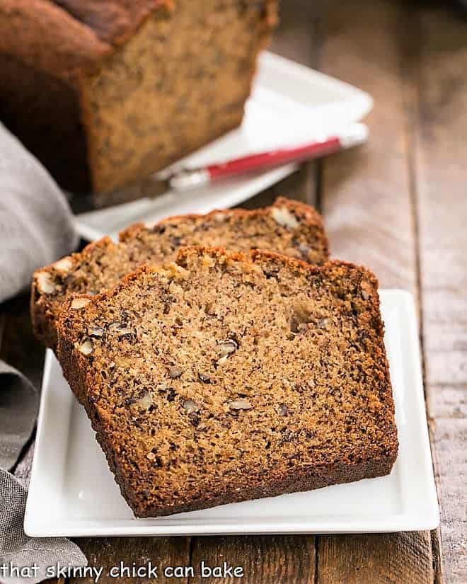 The Best Banana Nut Bread - Perfect brunch, breakfast or snack with the nutty flavor of both brown butter and pecans 2 slices of Best Banana Bread on a square white plate with the rest of the loaf in the background,