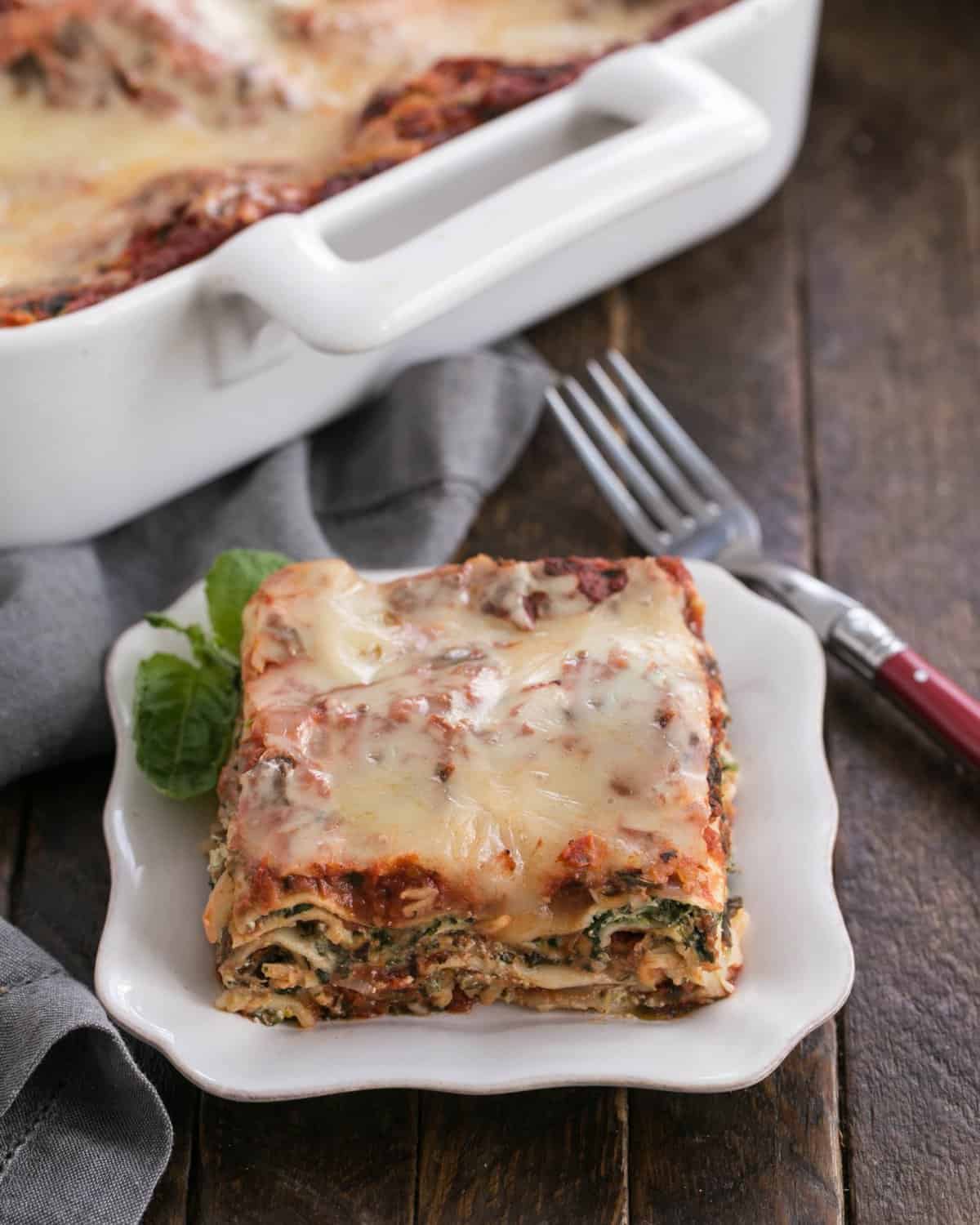 One slice of spinach lasagna on a white plate in front of the casserole dish.
