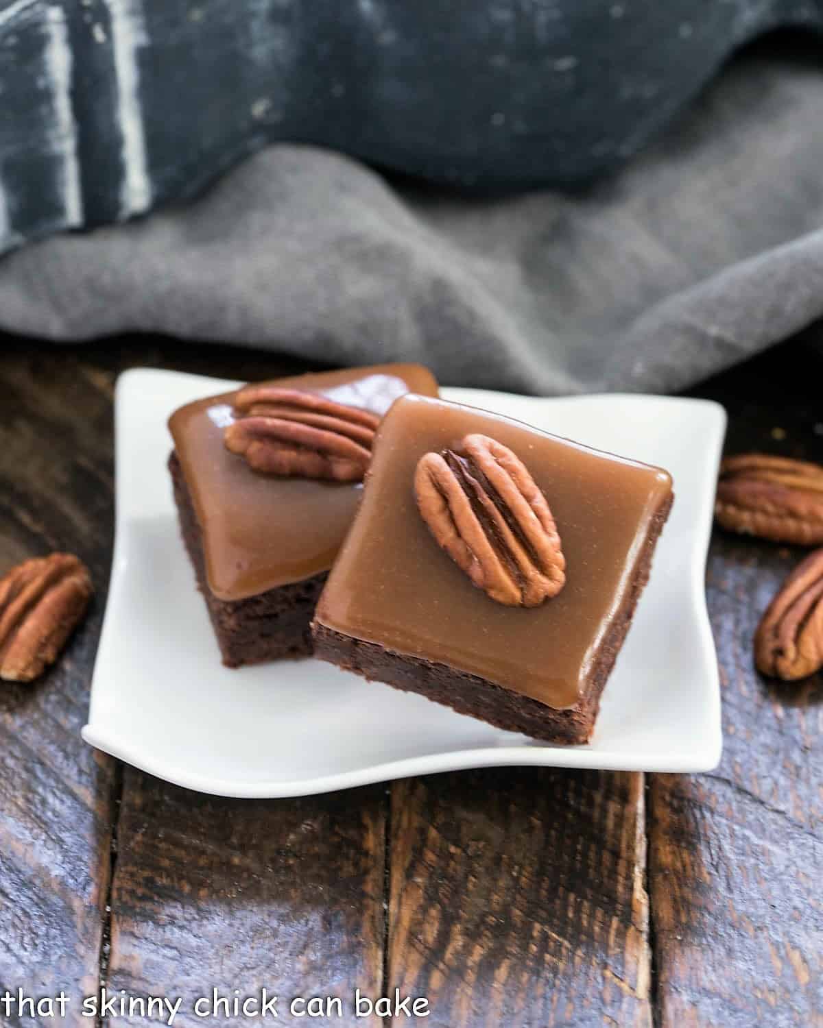 The Ultimate Turtle Brownies - fudgy brownies topped with the perfect chewy caramel and pecan halves Two turtle brownies on a small white wavy plate.