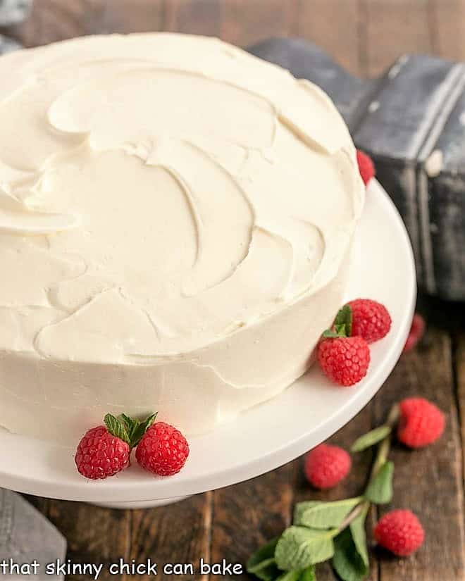 Vanilla Cake with White Chocolate Swiss Meringue Buttercream - a dense one layer vanilla cake topped with a luscious white chocolate frosting Close up of vanilla cae on a white cake stand wih raspberries and mint garnishes.
