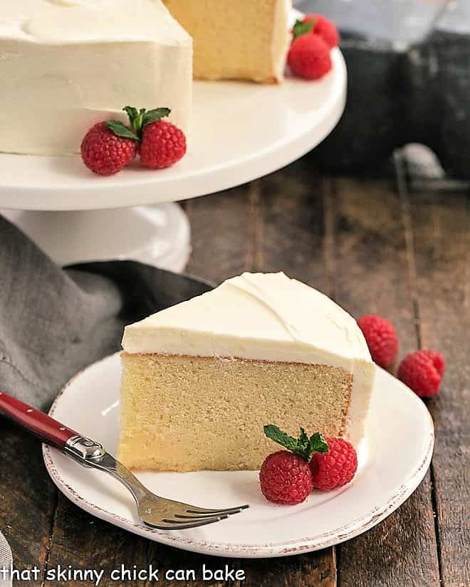 One Layer White Cake Recipe - a dense one layer vanilla cake topped with a luscious white chocolate buttercream Vanilla Cake with White Chocolate Buttercream on a white cake stand with one slice in the foreground.