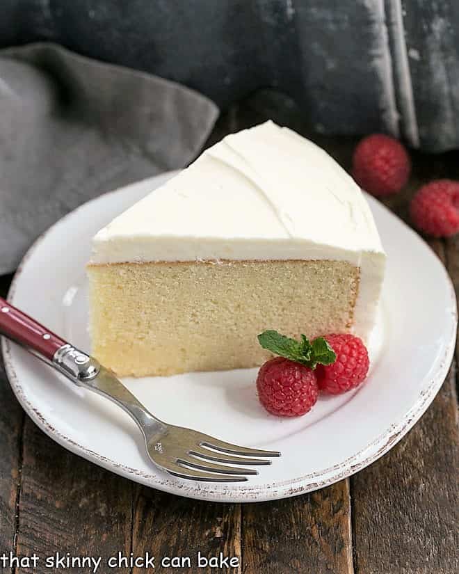 Vanilla Cake with White Chocolate Buttercream - a dense one layer vanilla cake topped with a luscious white chocolate buttercream, white chocolate curls and raspberries A slice of this white cake recipe on a white dessert plate with a red handled fork and raspberry garnish.