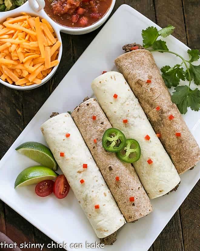 Soft Shell Tacos Recipe- An easy, homemade beef taco recipe that only takes a few more minutes than adding a packet of taco seasoning. Plus they're a whole lot tastier when they're homemade! Overhead view of 4 soft shell beef tacos on a white platter with peppers and cilantro to garnish.
