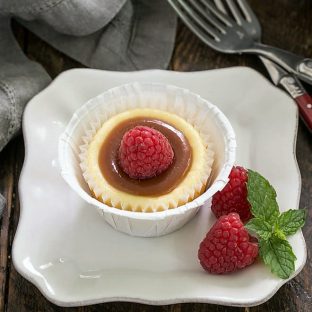 One Carame topped mini cheesecake on a square white plate with raspberries to garnish