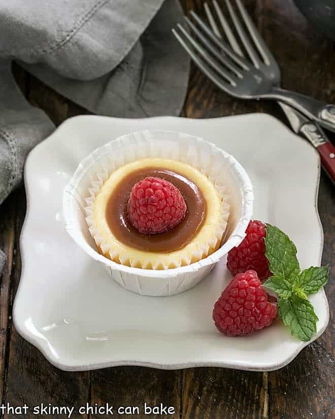 One Carame topped mini cheesecake on a square white plate with raspberries to garnish