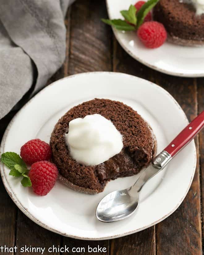 Overhead with of a chocolate lava cake topped with Kahlua whipped cream on a white plate with a raspberry and mint garnish and a red handle spoon.