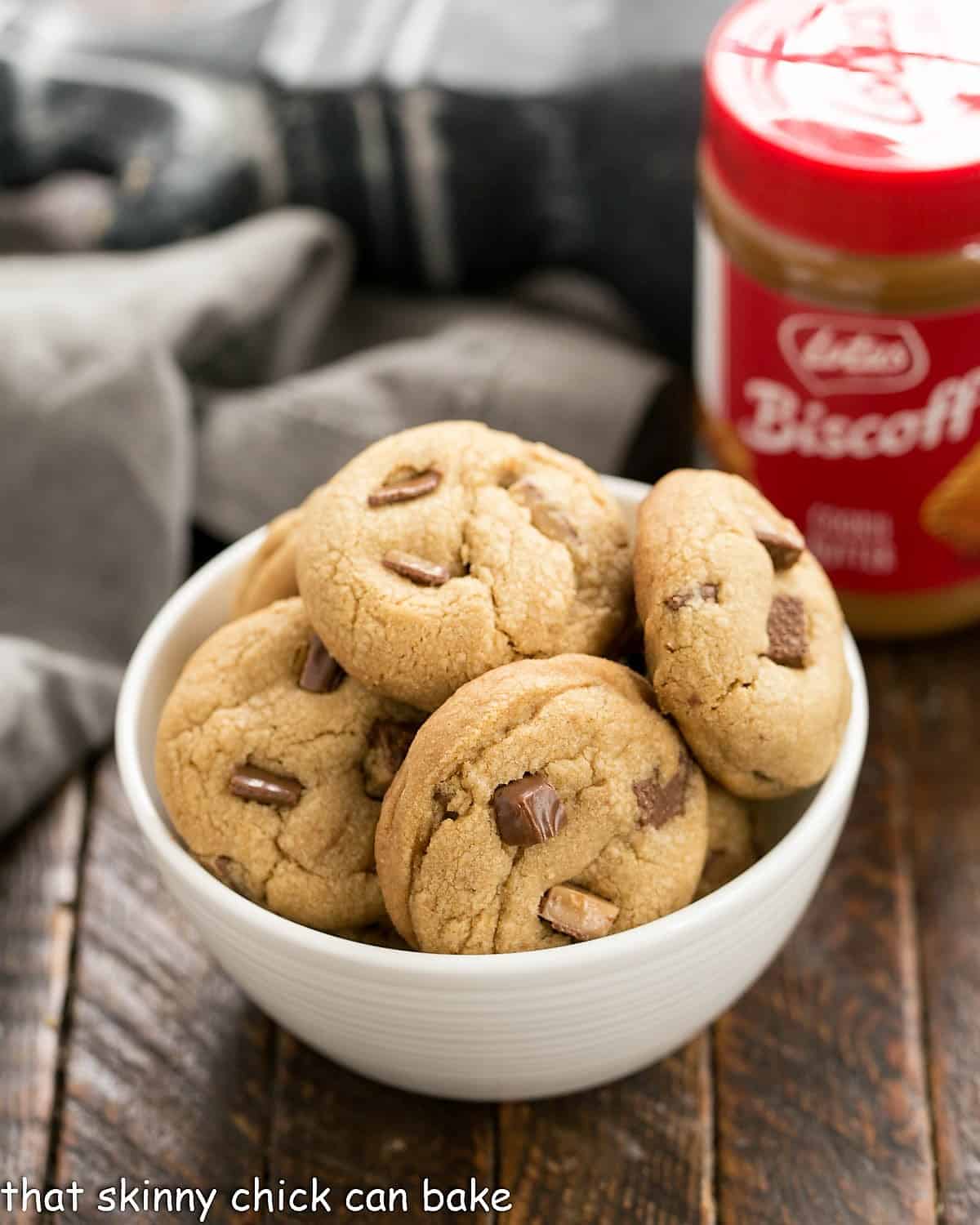 Biscoff Toffee Cookies - flavored with delicious cookie butter and enhanced with toffee chunks, these cookies are out of this world! a white bowl filled with biscoff cookies with a jar of biscoff butter in the backgroune