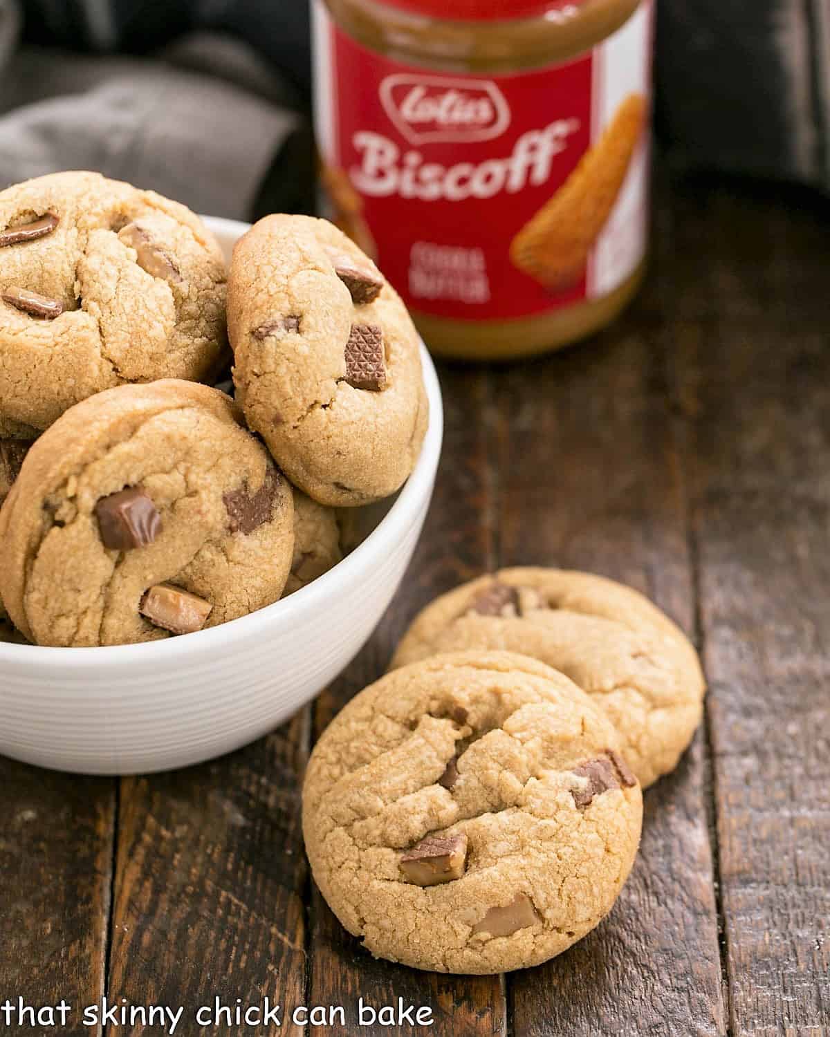 Two toffee biscoff cookies next to a white bowl of cookies and jar of cookie butter.