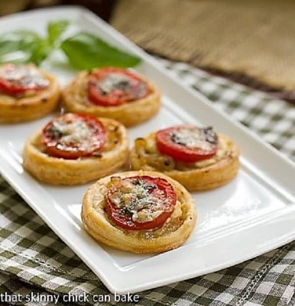 Tomato and Boursin Tartlets - an elegant appetizer with layers of flavor over puff pastry rounds Tomato and Boursin Tartlets lined up on a white ceramic tray with a sprig of basil.