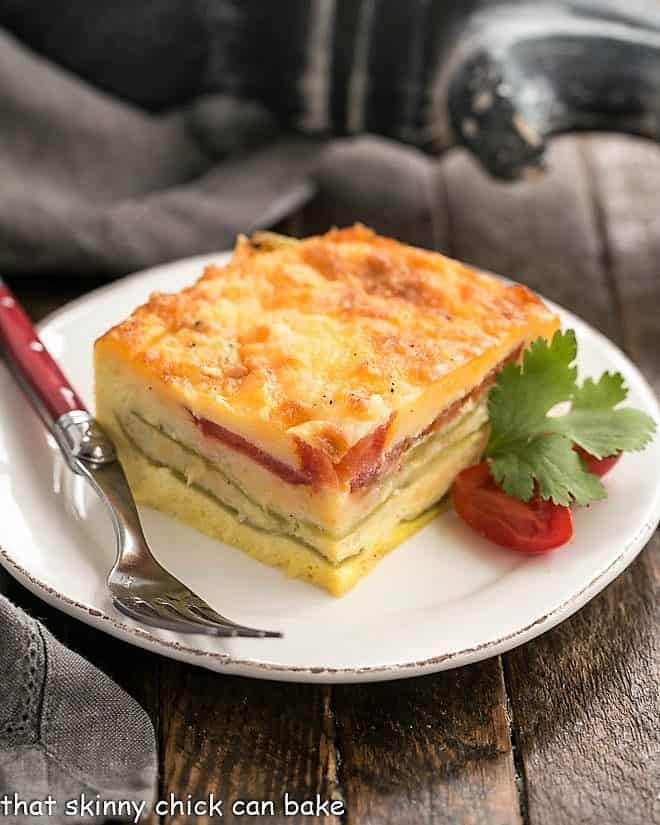 Chile Relleno Casserole - Layers of gooey cheese, eggs, tomatoes and peppers make for an irresistible Tex-Mex egg casserole dish! Chile Relleno Casserole slice on a white plate with cherry tomatoes and cilantro sprig.