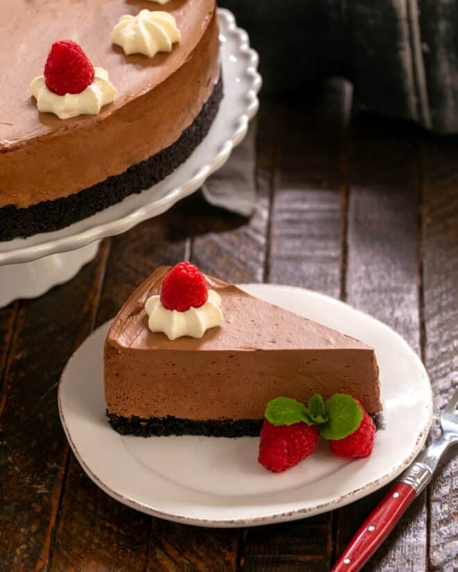 A slice of mousse cake on a white dessert plate in front of a cake stand.