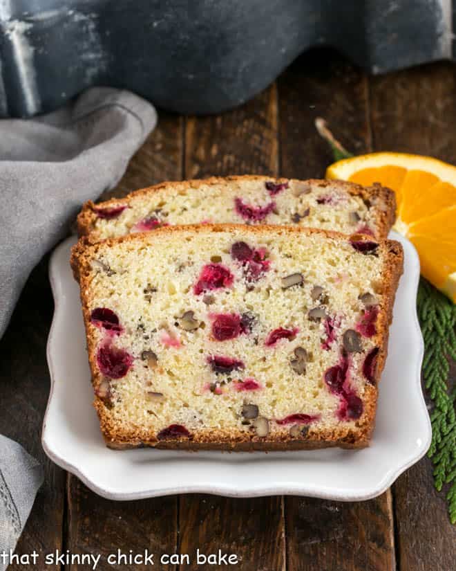 Classic Cranberry Pecan Bread is perfect for the holidays. The sweet-tart, crunchy-crispy components of this cranberry quick bread recipe make this a loaf to remember! 2 slices of cranberry bread on a white plate with an orange slice to garnish.
