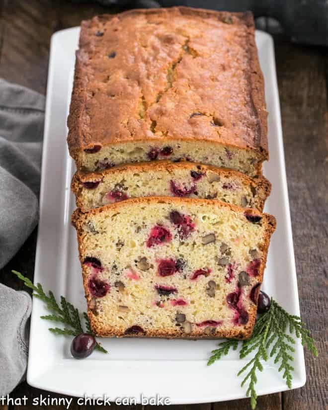 Classic Cranberry Pecan Bread is perfect for the holidays. The sweet-tart, crunchy-crispy components of this cranberry quick bread recipe make this a loaf to remember! Loaf of cranberry bread on a white tray with two slices in front of the loaf.
