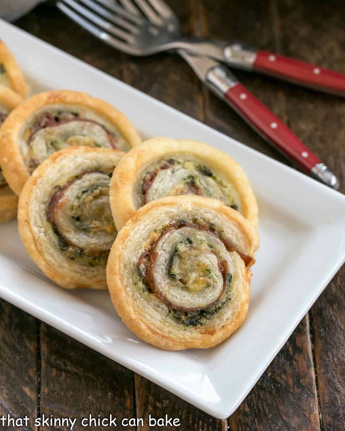 Close view of Prosciutto, Gruyere and Basil Pinwheels on a white ceramic tray with 2 forks.