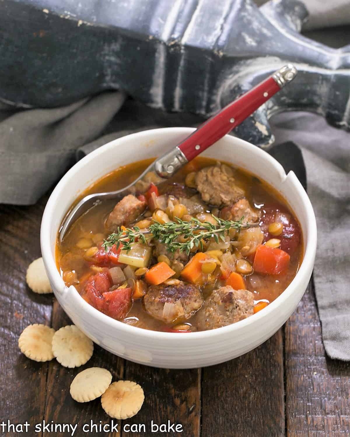 White bowl filled with lentil soup and a red handled spoon with oyster crackers.