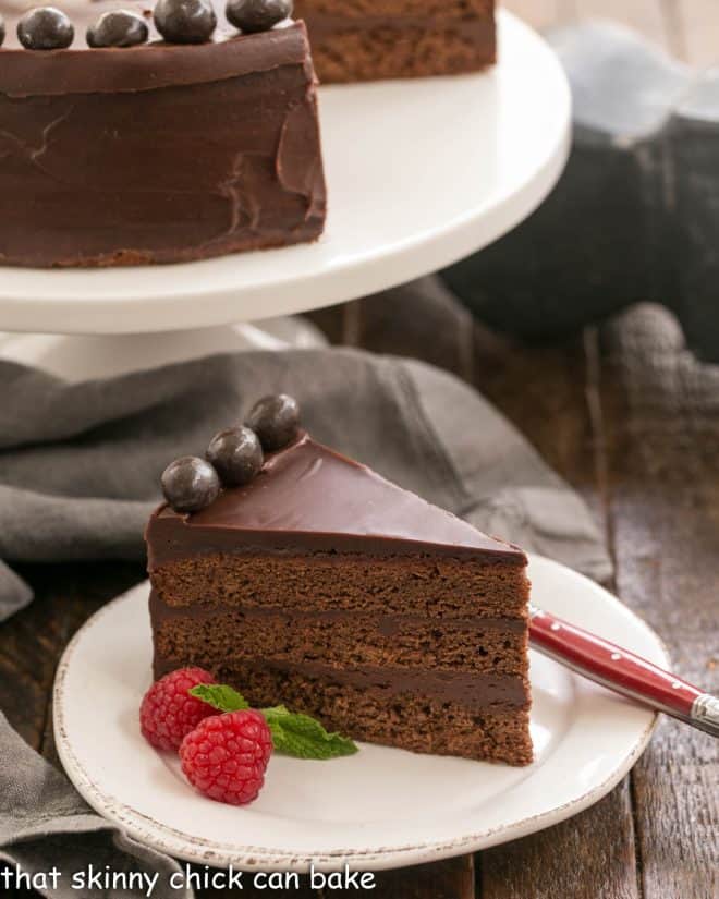 A slice of brownie cake in front of a white pedestal holding the rest of the cake