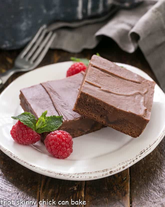 Chocolate Poke Brownies - The ganache seeps INTO the brownies!!! 2 poke brownies on a white dessert plate with raspberries and mint to garnish.