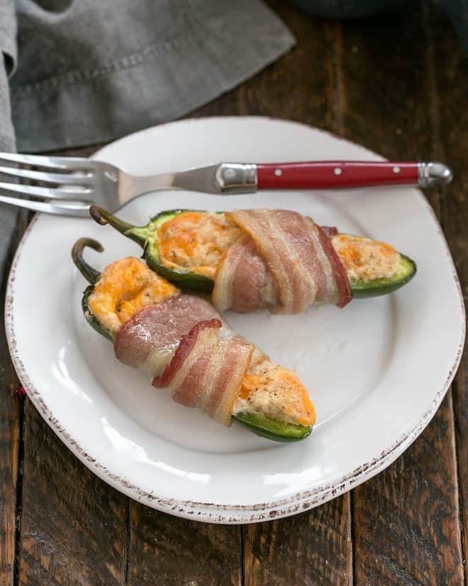 Jalapeno Poppers - filled with cream cheese and cheese before being wrapped in bacon, these appetizers will fly off the serving tray! Two bacon wrapped jalapeno poppers on a round white plate with a red handle fork.