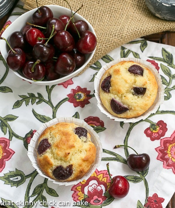 Cherry Buttermilk Muffins - Scrumptious, tender muffins with two kinds of cherries! Double Cherry Muffins on a floral napkin with a bowl of dark cherries in a white bowl.