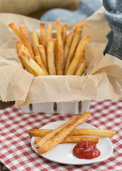 Thin Crispy French Fries in a basket on a checkered napkin with a plate with a few fries and a blob of ketchup.