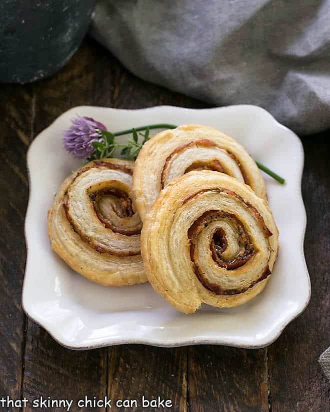 Honey Mustard Pepperoni Pinwheels - Puff pastry, mustard, Asiago and pepperoni are rolled into these irresistible pinwheels Honey Mustard Pepperoni Pinwheels on a square white plate garnished with flowering chives.