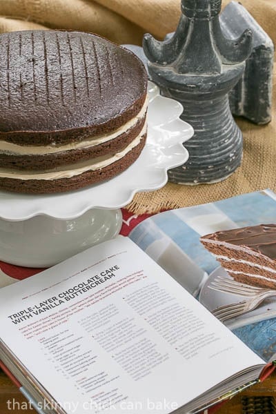  Chocolate Cake with Vanilla Buttercream Filling unfrosted on a cake stand next to an open cookbook