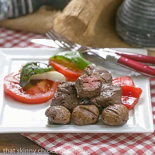 Tender Grilled Beef Steak Kabobs removed from their skewers on a square white plate with tomato, mushrooms and pepper garnishes. Tender Beef Steak Kabobs on a white plate