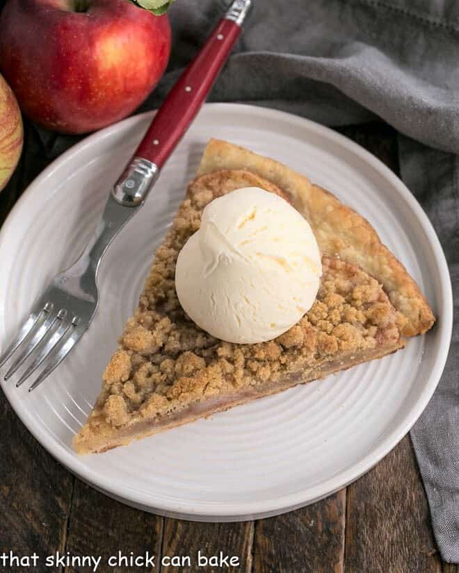 Apple Pie Pizza - A dessert pizza made with a pastry crust, spiced apples, and a crumb topping! An easy apple recipe with the taste of a Dutch Apple Pie! A slice of apple pie pizza on a white plate topped with a scoop of ice cream.