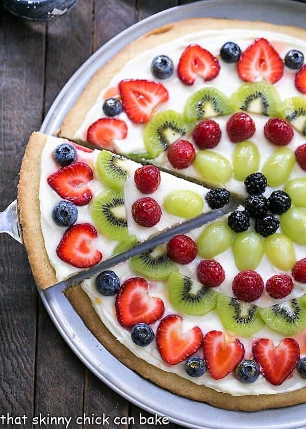 Fruit Pizza Dessert - A vibrant, delicious dessert pizza with a sugar cookie crust and cream cheese frosting! Overhead view of fresh fruit pizza with a slice being removed.