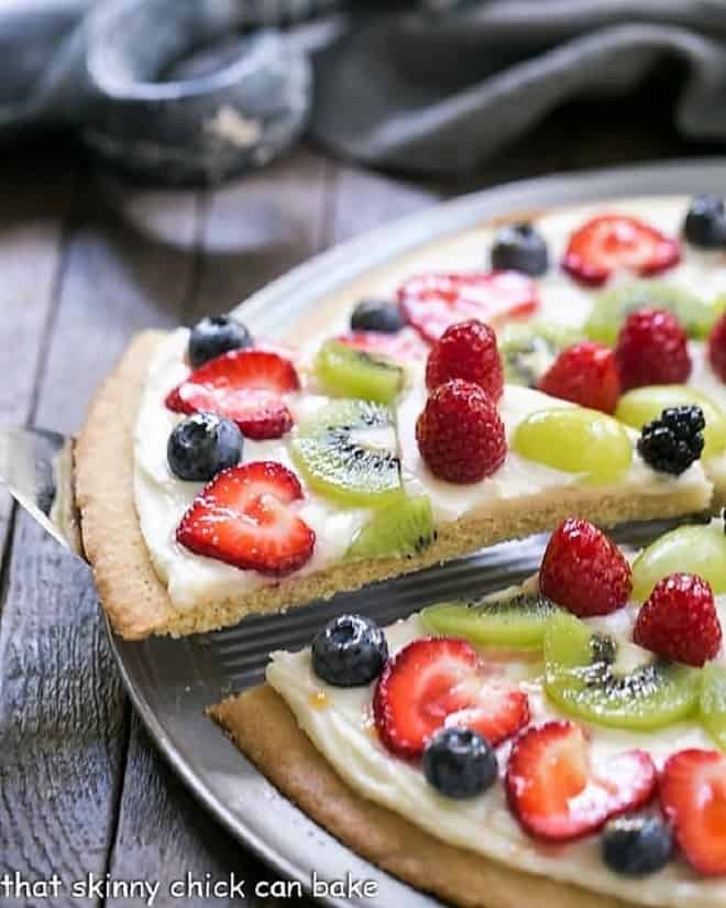 Fresh Fruit Pizza - A vibrant, delicious dessert pizza with a sugar cookie crust and cream cheese frosting! A slice of Fresh Fruit Pizza being removed from a whole pizza on a metal pizza pan.