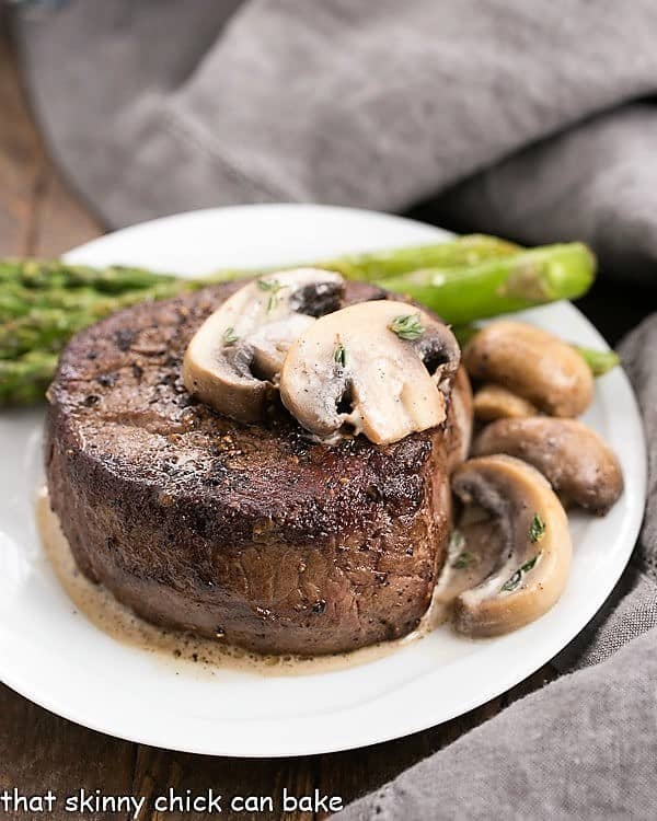 Mushroom Topped Pepper Steak topped with mushrooms on a white plate with asparagus spears.