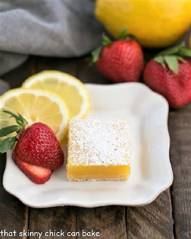 The Best Lemon Bars with a dreamy citrus layer over a sweet shortbread crust White dessert plate with the Best Lemon Bars, garnished with fresh strawberry slices and slices of lemon.