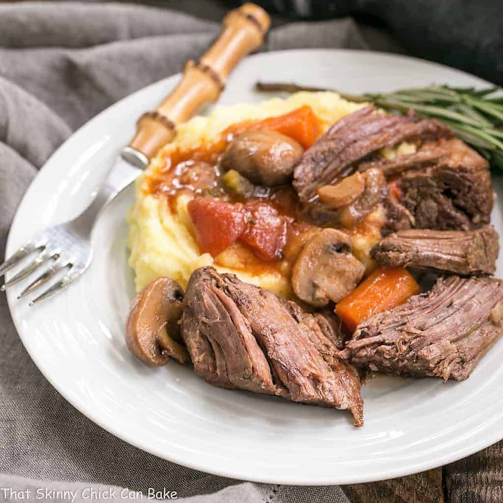 Pot Roast in oven Recipe - This is one unforgettable beef pot roast with an amazing depth of flavor! Pot Roast in Oven with Mushrooms, Tomatoes & Red Wine on a white ceramic plate with a bamboo handled fork.