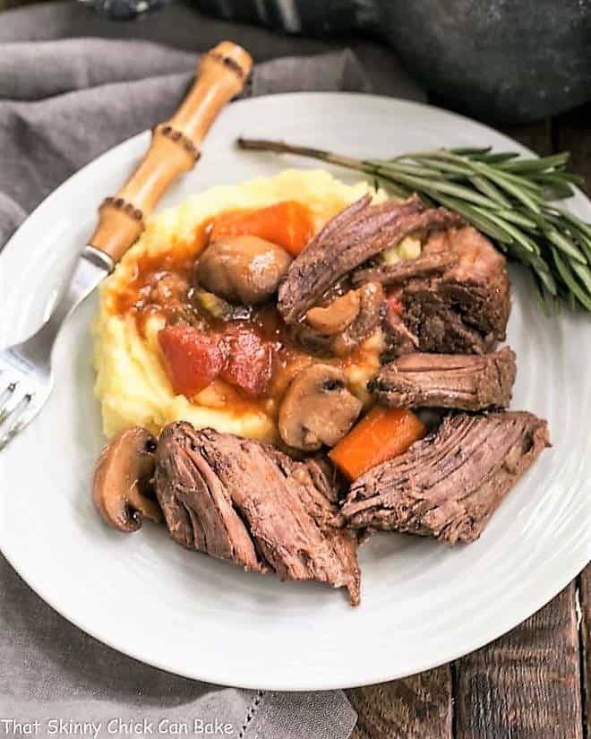 Oven Pot Roast Recipe with Mushrooms, Tomatoes & Red Wine - marvelous comfort food with an exquisite depth of flavor! Pot Roast with Mushrooms, Tomatoes & Red Wine on a white plate with mashed potatoes and a sprig of rosemary.