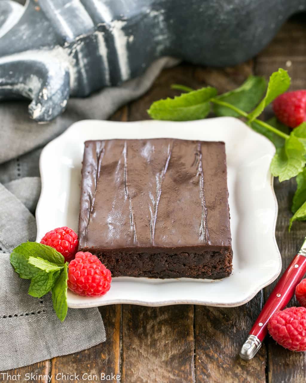 Slice of Fudgy Chocolate Sheet Cake with Cocoa Icing on a square white plate with raspberries and mint.
