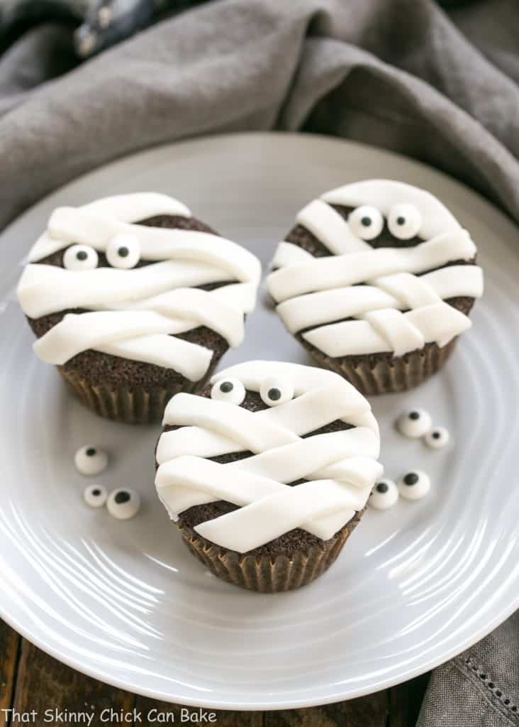 EASY Chocolate Mummy Cupcakes - A simple way to make festive Halloween cupcakes EASY Chocolate Mummy Cupcakes on a round white ceramic plate garnished with sugar eyes