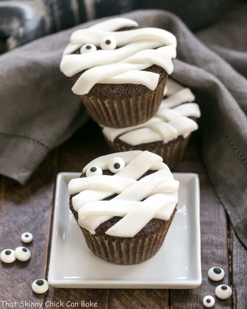 EASY Chocolate Mummy Cupcakes - A simple way to make festive Halloween cupcakes EASY Chocolate Mummy Cupcakes on a white plate and stacked with sugar eyes to garnish