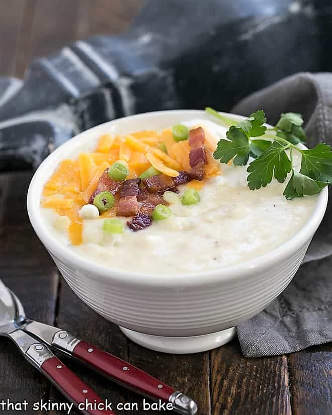 Loaded Baked Potato Soup - A creamy easy potato soup with chunks of baked potatoes and topped with cheddar, bacon, sour cream and chives! Side view of Loaded Baked Potato Soup in a white ceramic soup bowl with two red handled soup spoons.
