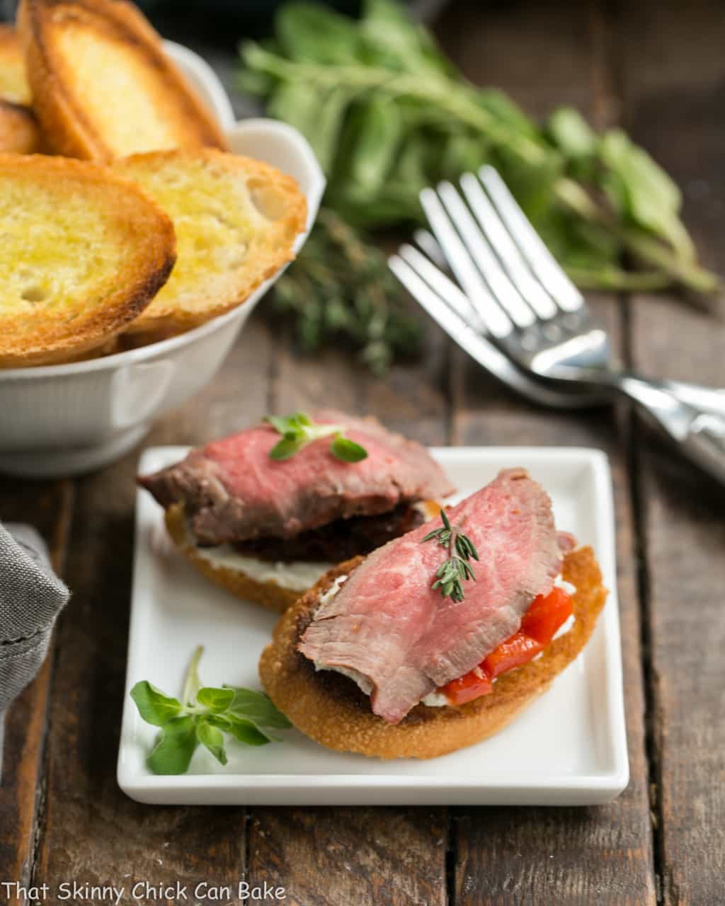 Beef Crostini with Blue Cheese - Baguette toasts smeared with a blue cheese spread and topped with peppers or tomatoes and rare grilled beef! Beef Crostini with Blue Cheese on a square white appetizer plate.