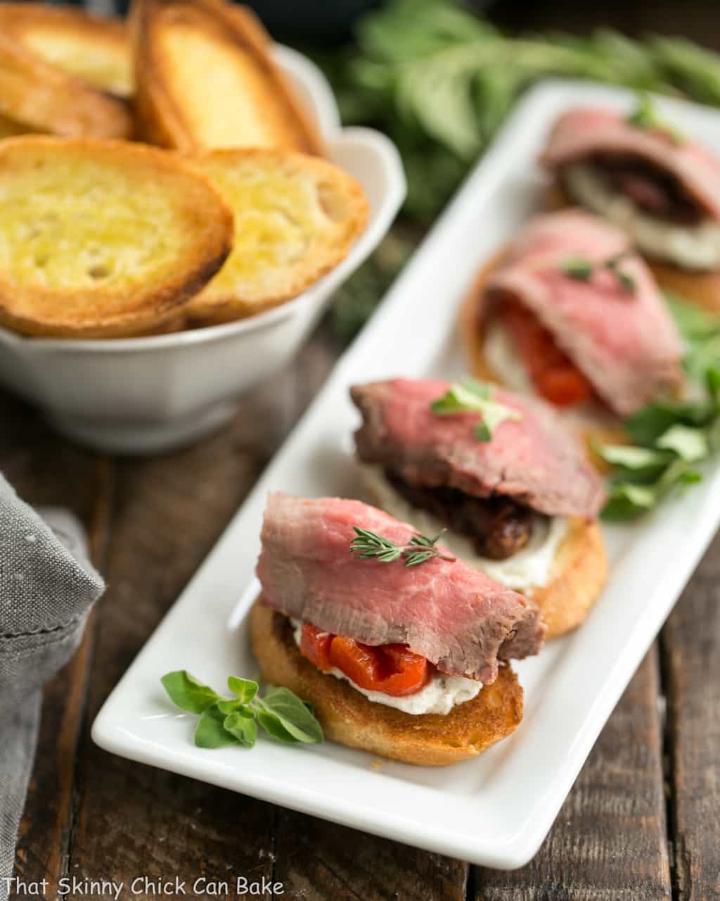 Roast beef appetizer idea - Baguette toasts smeared with a blue cheese spread and topped with peppers or tomatoes and rare grilled beef! Beef Crostini with Blue Cheese Spread lined up on a white platter.