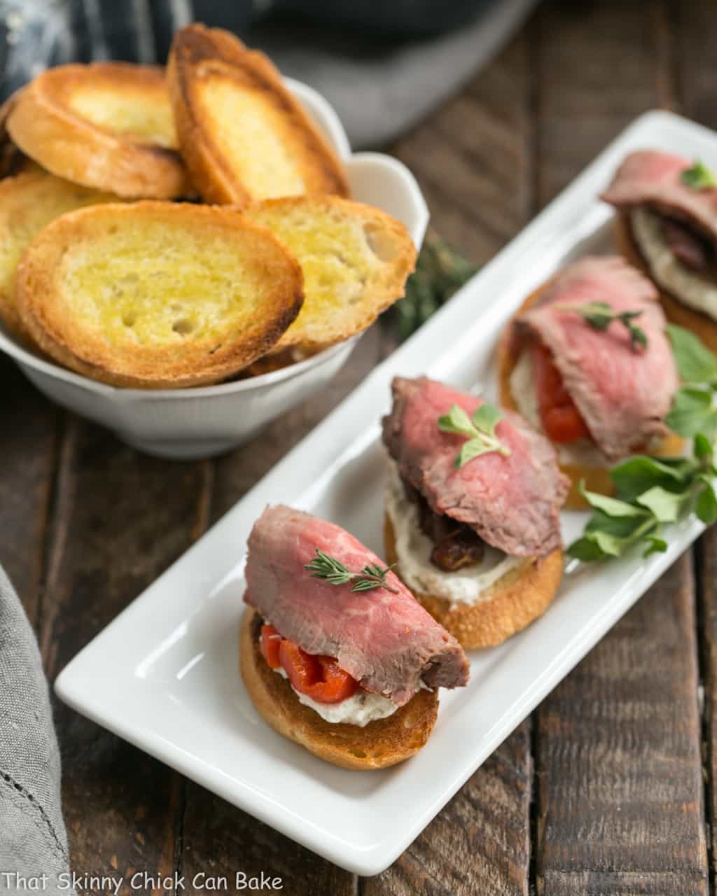 Beef Brusctetta Recipe with Blue Cheese - Baguette toasts smeared with a blue cheese spread and topped with peppers or tomatoes and rare grilled beef! Beef Crostini with Blue Cheese Spread on a narrow white tray viewed from above.