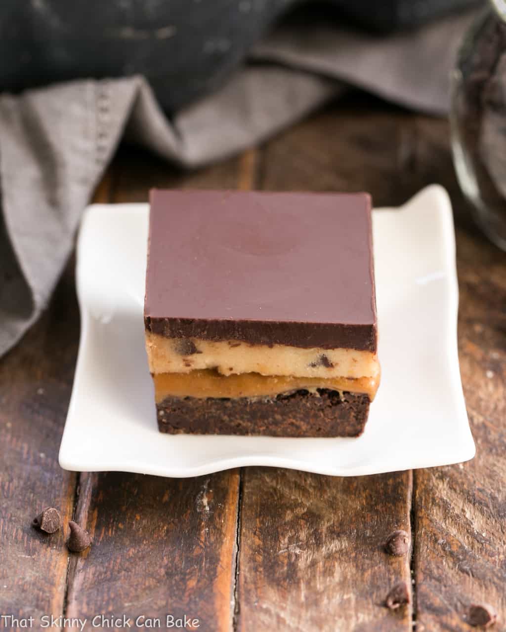 Cookie Dough Billionaire Bars - Layers of brownies, caramel, cookie dough and ganache for an outrageously rich dessert! Cookie Dough Billionaire Bars on a small, wavy white plate.