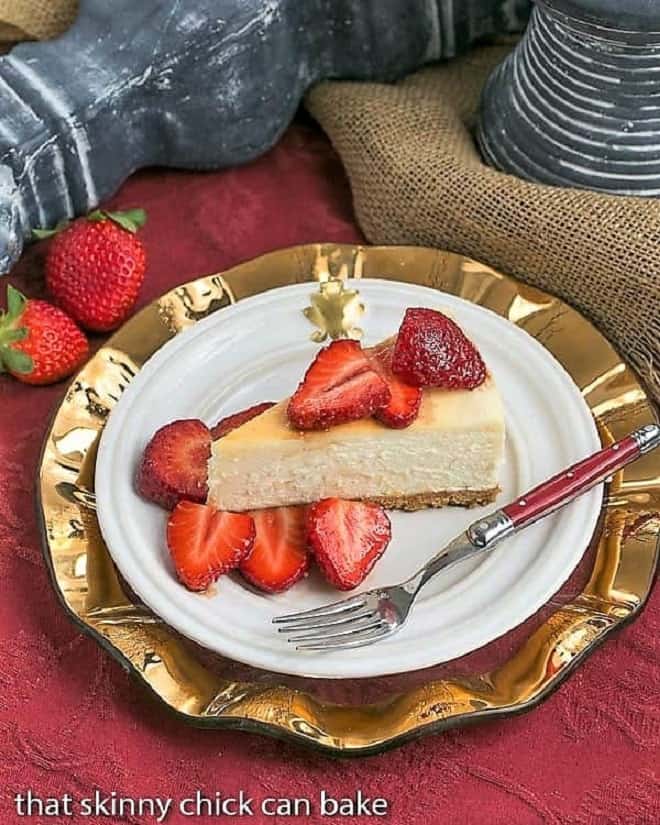 Homemade Cheesecake with Mascarpone Balsamic Strawberries - The most creamy, luscious cheesecake topped with sweetened balsamic strawberries A slice of Mascarpone Cheesecake with Balsamic Strawberries on a white plate on top of a gold plate with a red handled fork.